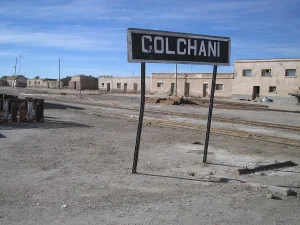 Colchani station sign above railroad tracks with weathered adobe buildings in the background under a blue sky