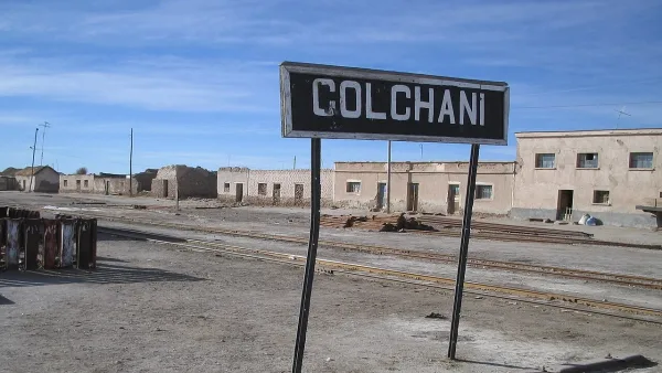Letrero de estación ferroviaria que dice Colchani, con vías de tren y edificios de adobe en mal estado al fondo.