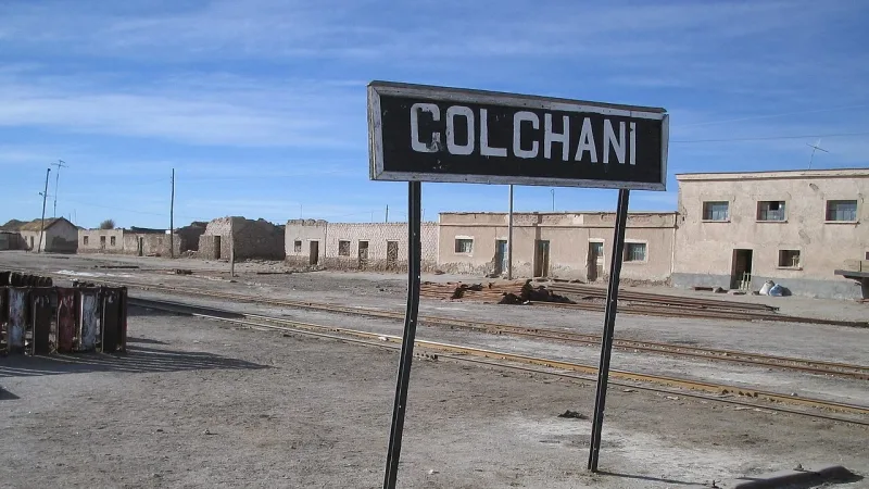 Colchani station sign above railroad tracks with weathered adobe buildings in the background under a blue sky