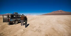 Dos personas junto a una camioneta 4x4 en el desierto de Chiguana, con volcanes y cielo azul al fondo.