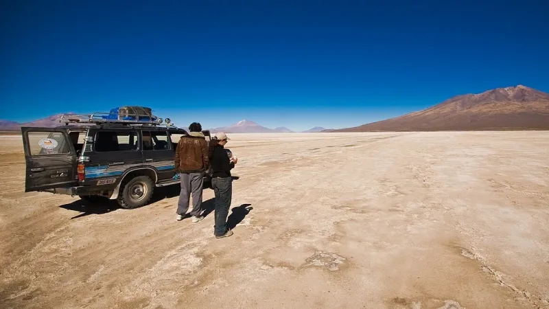 Dos personas junto a una camioneta 4x4 en el desierto de Chiguana, con volcanes y cielo azul al fondo.