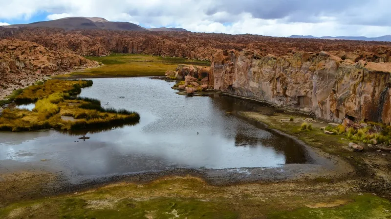 Laguna Catal cercada por rochas vulcânicas avermelhadas, vegetação rasteira e reflexo de nuvens na água calma do altiplano