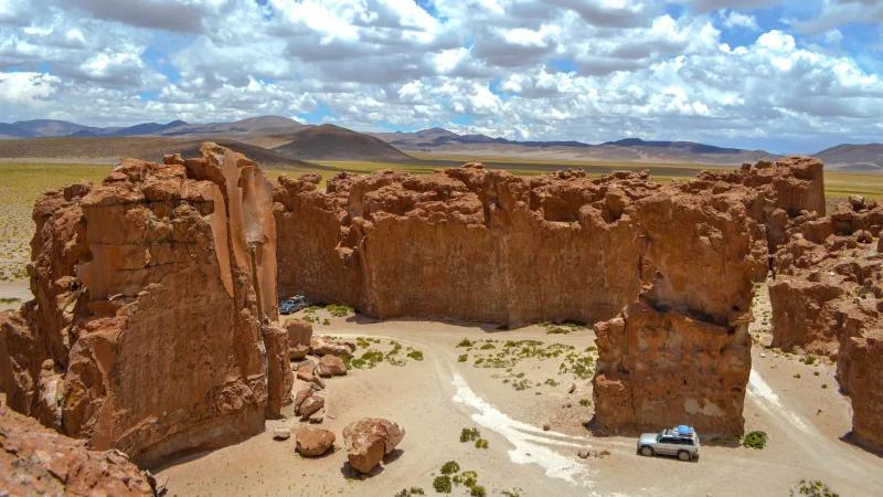 Ruínas de pedra vermelha no altiplano boliviano com dois veículos 4x4 ao redor, planície e montanhas ao fundo