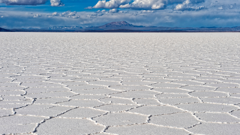 Vasta planície branca do Salar de Uyuni com padrões hexagonais de sal, montanhas ao fundo e céu azul com nuvens.