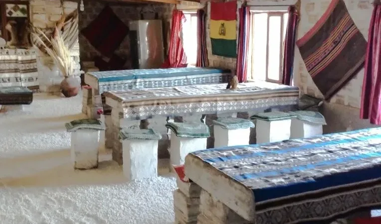 Interior de hotel de sal en el Salar de Uyuni con mesas y sillas de sal, bandera boliviana y textiles andinos.