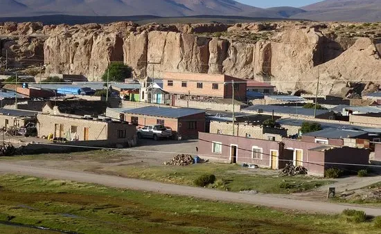 Pequena vila andina com casas simples, formações rochosas e montanhas ao fundo sob céu azul claro