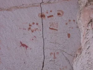 Pinturas rupestres em tons vermelhos na rocha, com figuras humanas, animais e símbolos abstratos em parede de pedra rachada.