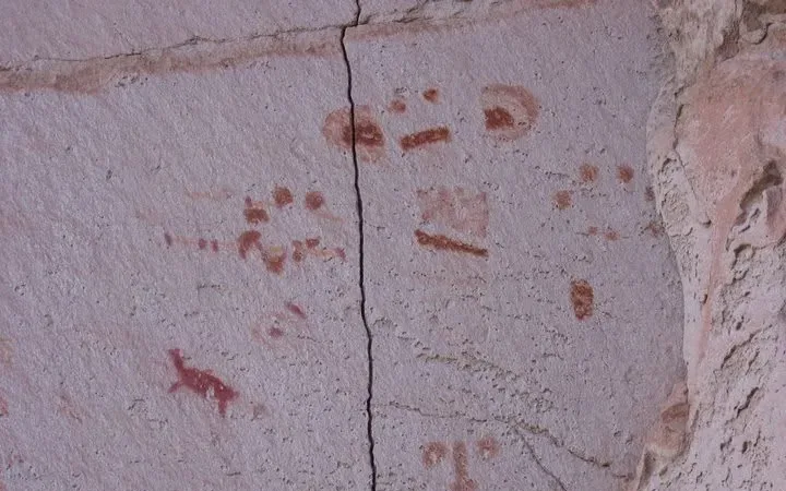 Pinturas rupestres em tons vermelhos na rocha, com figuras humanas, animais e símbolos abstratos em parede de pedra rachada.