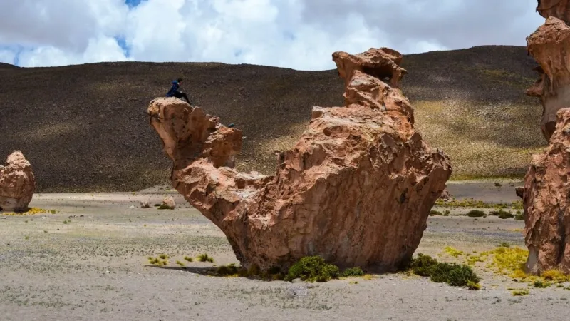 Formación rocosa erosionada con forma de camello en el altiplano boliviano, con una persona sentada sobre la roca.